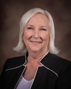 A woman with short white hair and a black jacket smiles against a dark, neutral background.