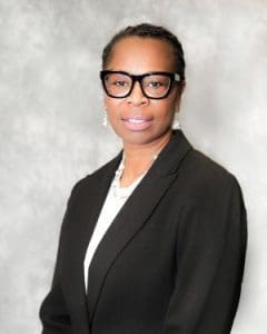 A person wearing glasses and a black blazer stands against a gray background.