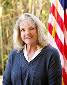 A woman with blonde hair wearing a dark sweater and white collared shirt stands in front of an American flag and greenery.